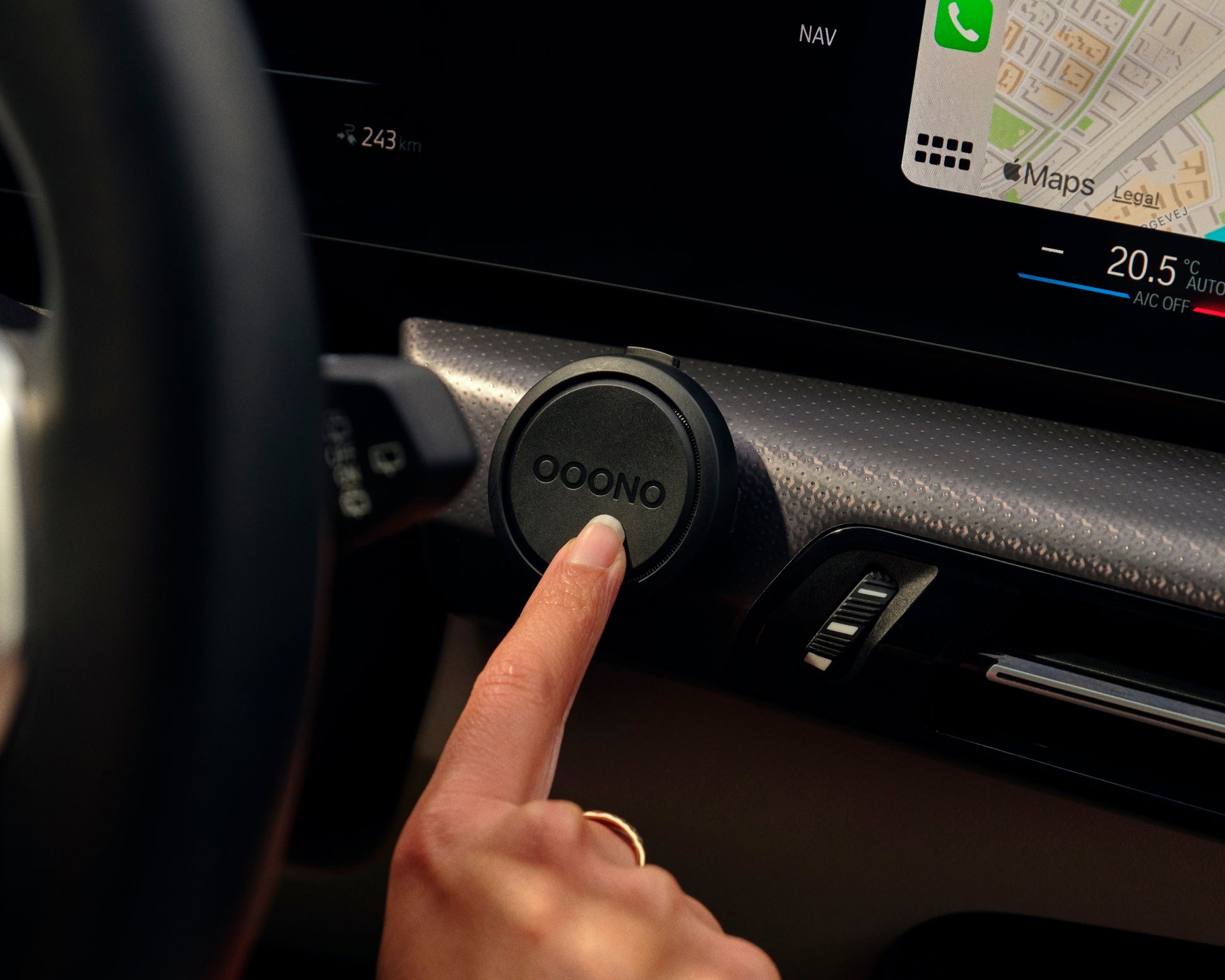 A hand with a ring presses a round black speed camera detector labeled Ooono, mounted on a car dashboard, with a GPS map and temperature display visible on the car’s screen.